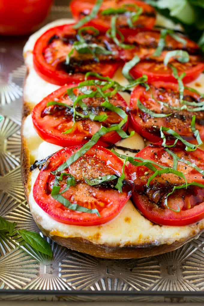 A split loaf of caprese bread on a sheet pan topped with fresh mozzarella cheese, sliced red tomatoes, thinly sliced basil and balsamic glaze.