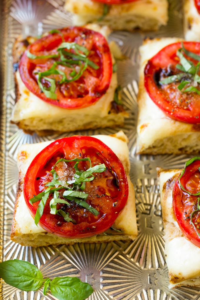 Bread cut into squares topped with melted mozzarella cheese, tomato slices, basil and balsamic glaze.