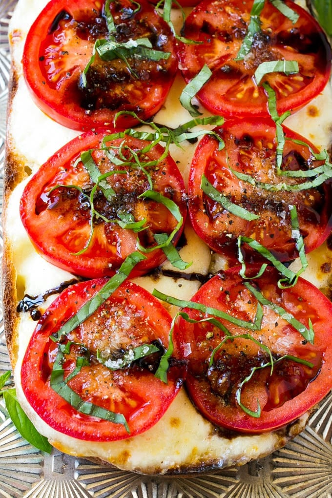 A loaf of bread cut in half, topped with melted mozzarella cheese, tomato slices, chopped basil and balsamic glaze.