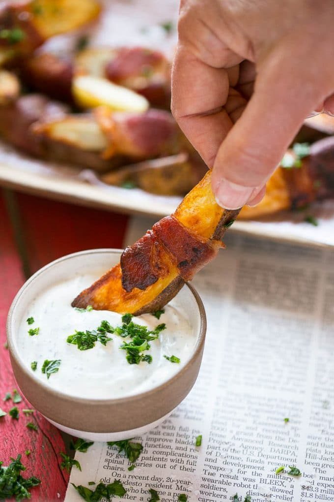 A hand dipping a bacon wrapped potato wedge into ranch.