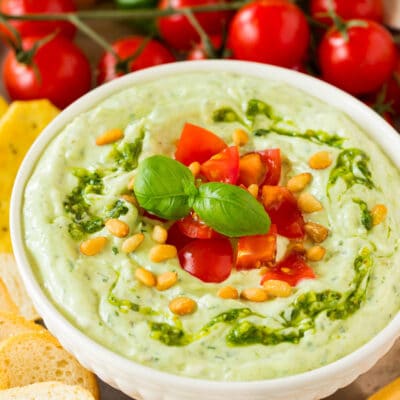 A bowl of pesto dipped topped with tomatoes and served with breads and vegetables.