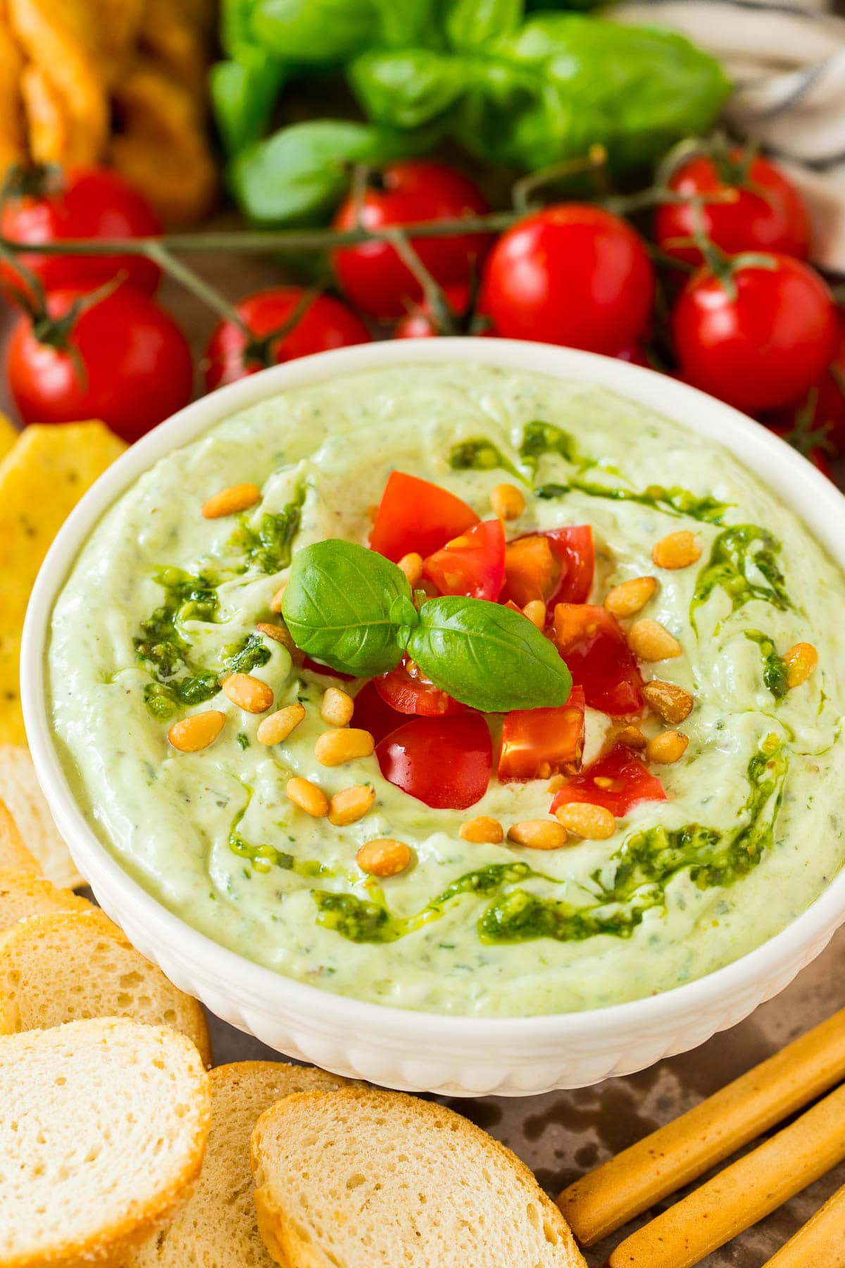 A bowl of pesto dipped topped with tomatoes and served with breads and vegetables.