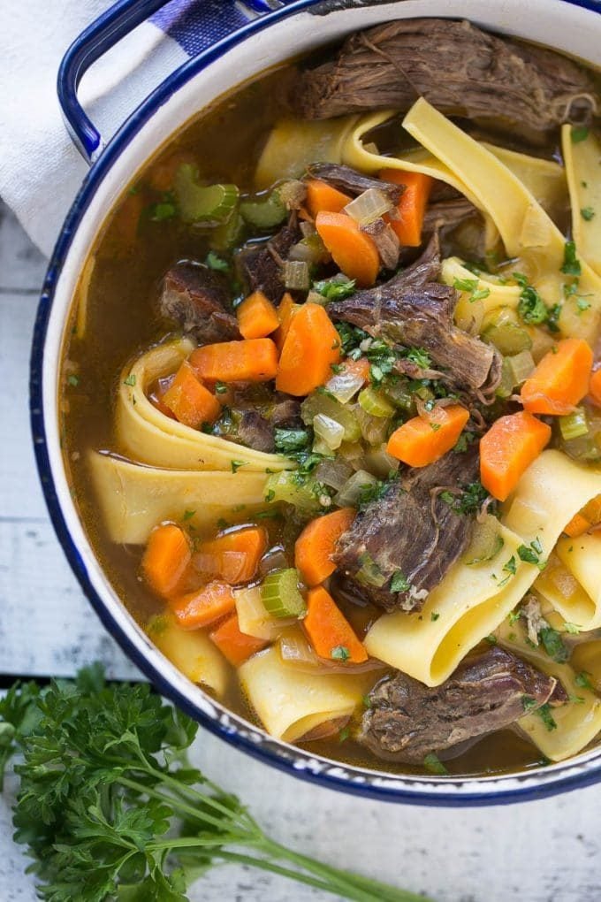 A pot full of beef soup with egg noodles, carrots, celery and parsley.