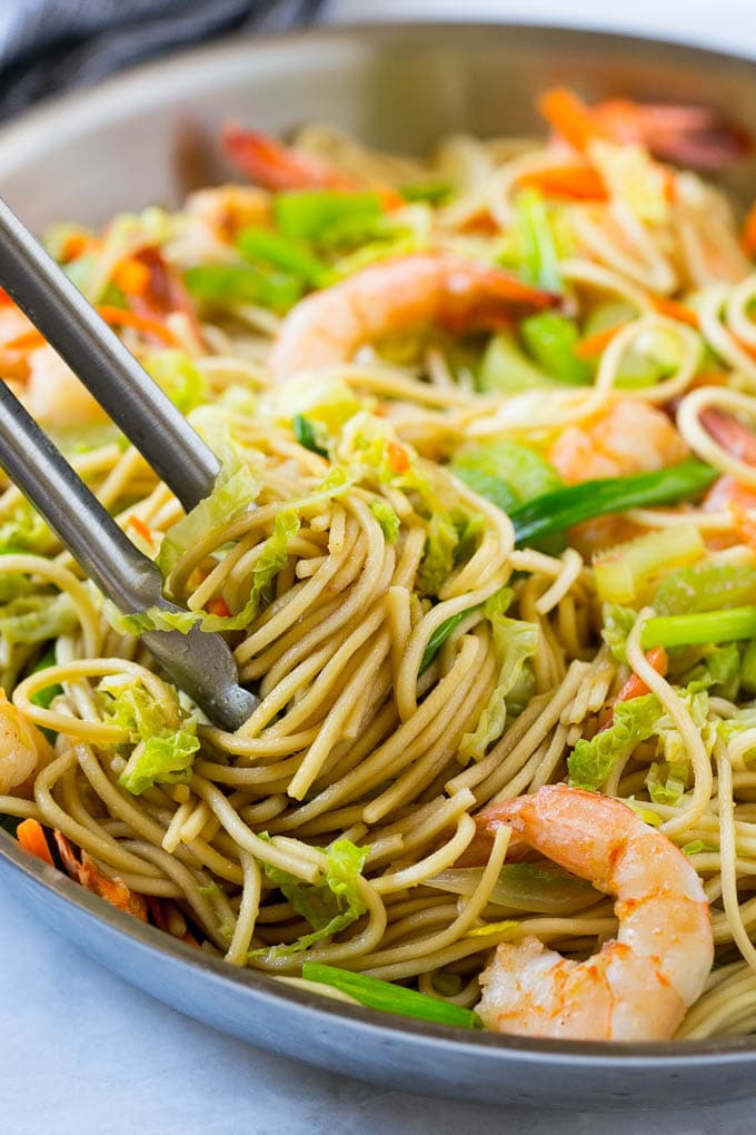 A pan filled with shrimp chow mein with a pair of tongs lifting out the noodles.