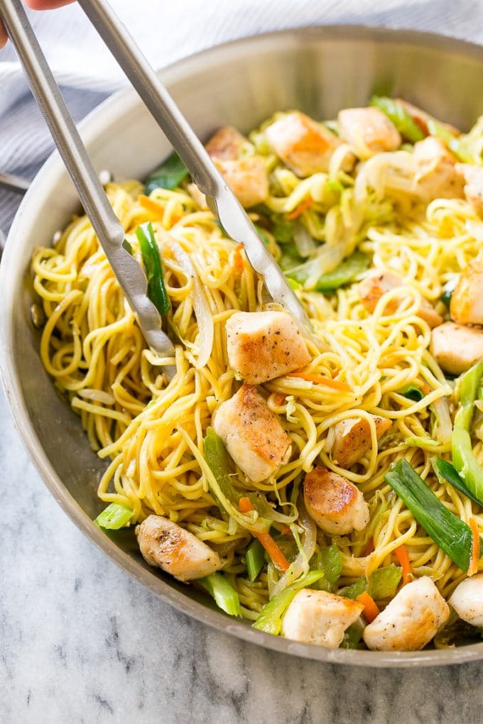 A pan of chicken chow mein with tongs pulling out a serving.