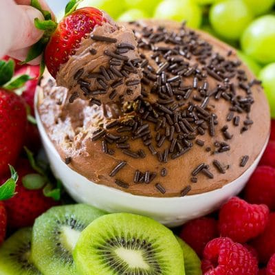 A strawberry in a bowl of chocolate fruit dip, surrounded by assorted fruit.