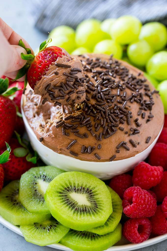 A strawberry in a bowl of chocolate fruit dip, surrounded by assorted fruit.