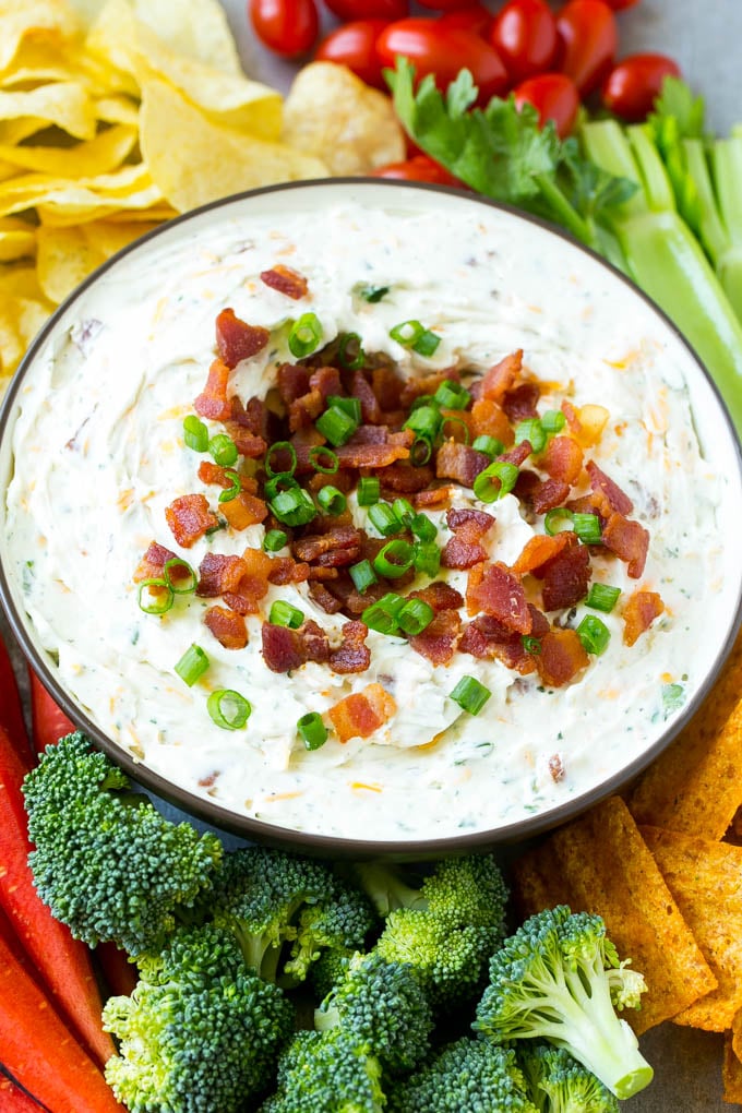 A bowl of creamy bacon cheddar ranch dip surrounded by vegetables and chips.