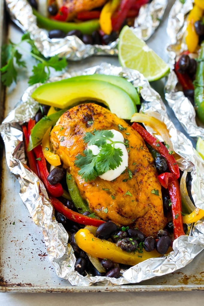 Cooked chicken breasts covered in cheese surrounded by peppers, onions and black beans, all inside a foil packet.