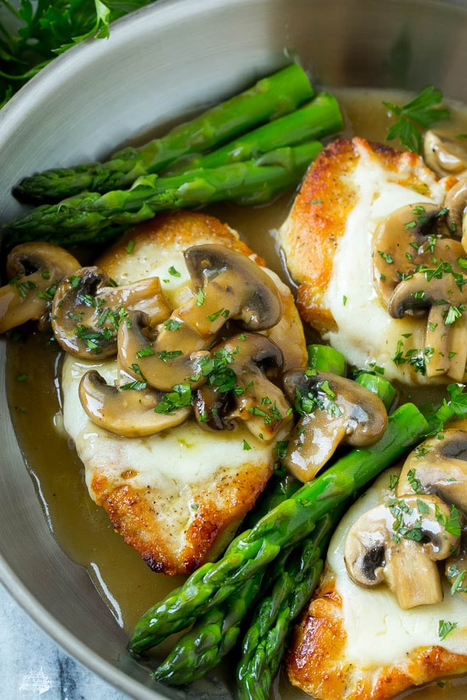 Golden brown chicken breasts topped with melted mozzarella cheese and mushroom sauce, in a pan with asparagus.