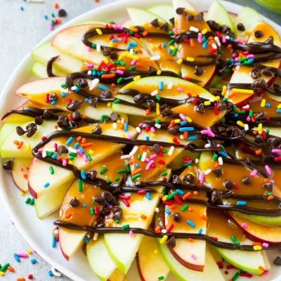 A plate of sliced apples topped with caramel and chocolate sauces, rainbow sprinkles and mini chocolate chips
