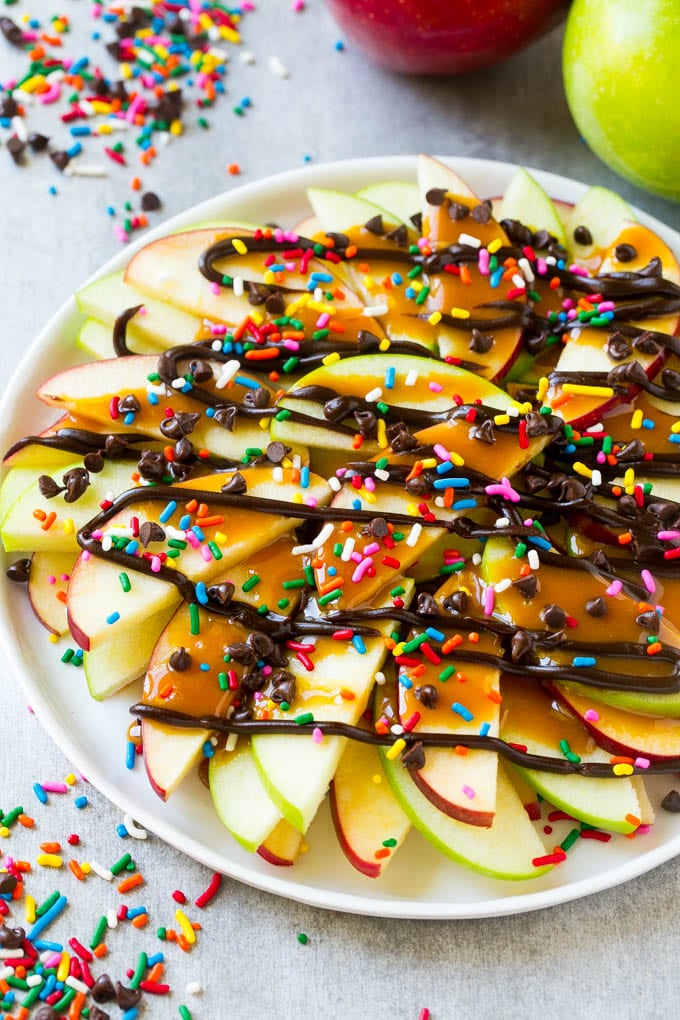 A plate of sliced apple nachos topped with caramel and chocolate sauces, rainbow sprinkles and mini chocolate chips