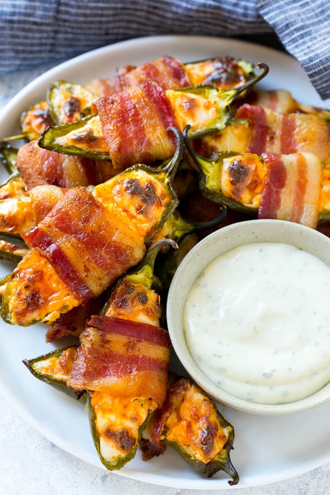 A plate full of bacon wrapped jalapeno poppers with a side of ranch for dipping.