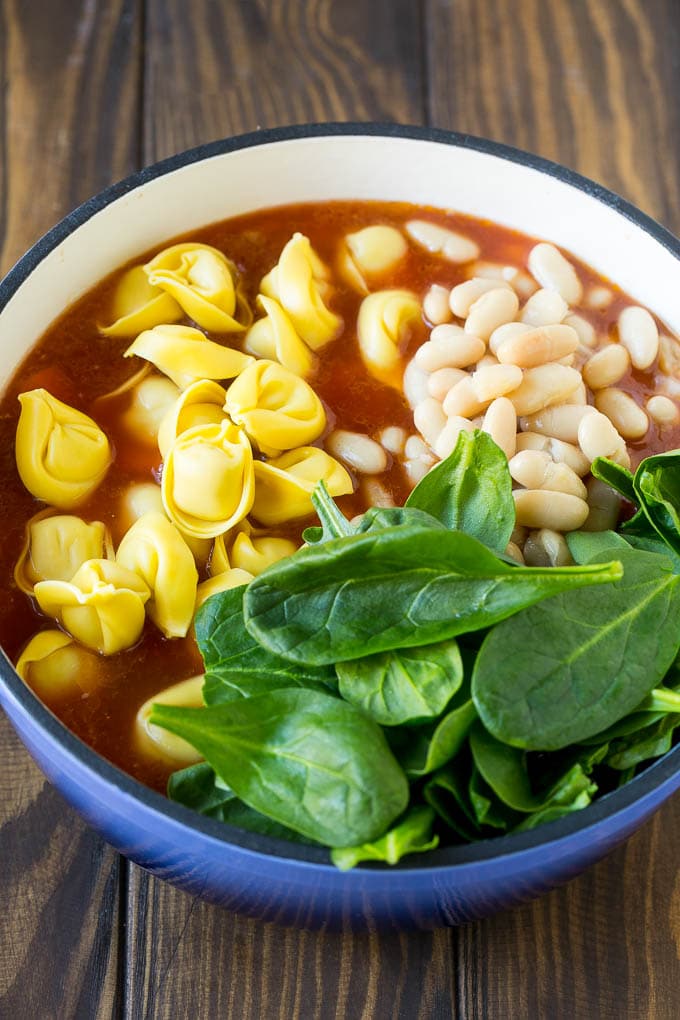 A pot of soup with tortellini, spinach leaves and white beans.