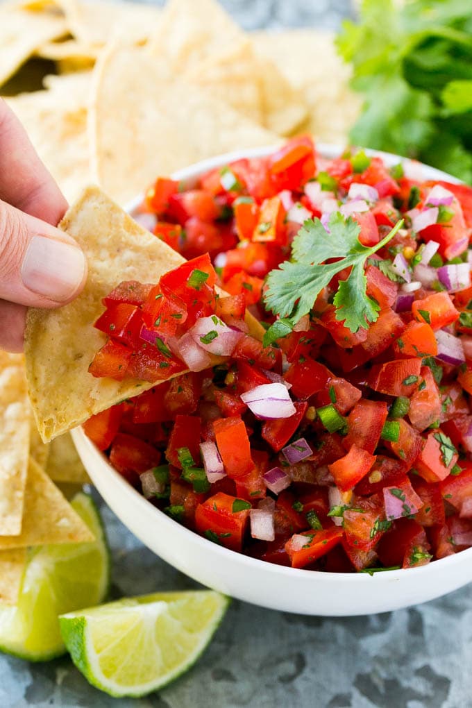 A hand with a chip taking a scoop of pico de gallo from a bowl.