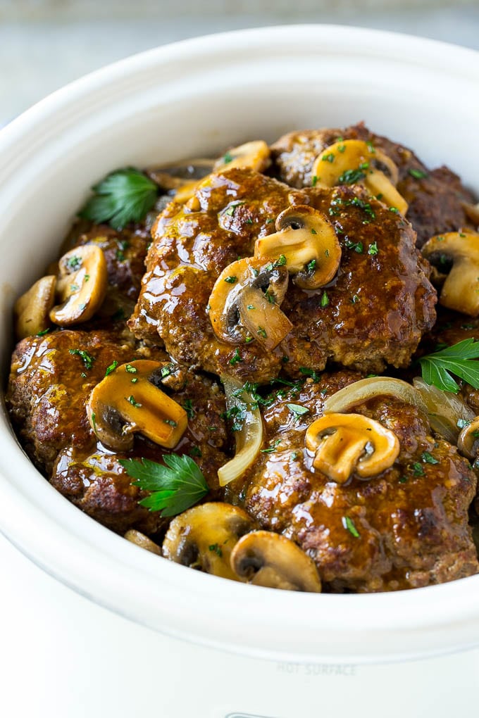 A crock pot with hamburger patties, cooked mushrooms and gravy.