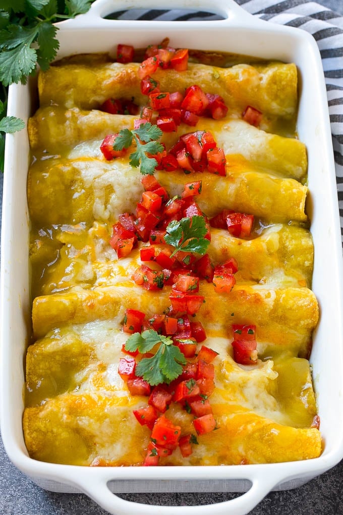 A baking dish of green chile chicken enchiladas topped with cheese, tomatoes and cilantro leaves.