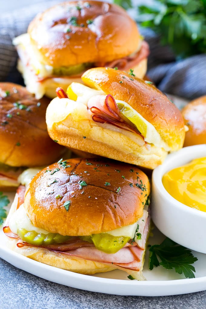 A plate full of Cuban sliders with cheese, pickles and ham on buns.
