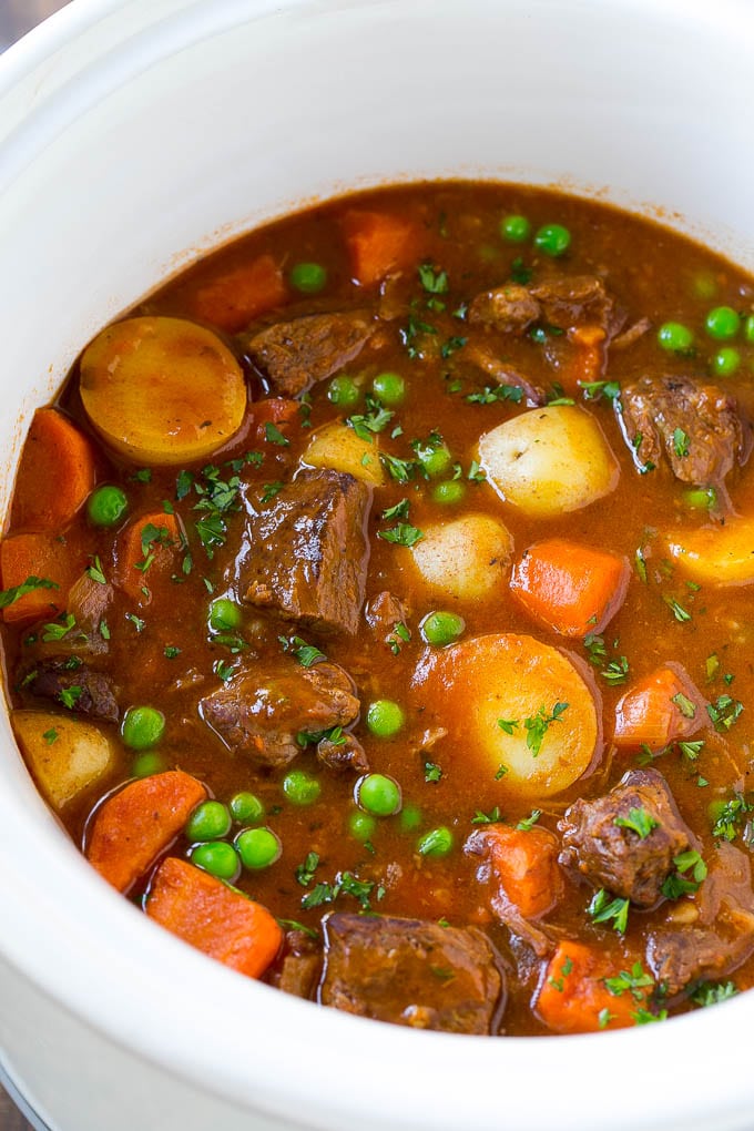 A slow cooker full of beef stew that contains meat, potatoes, carrots and peas, all in a savory broth.