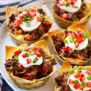 A plate of taco cups made with wontons, seasoned ground beef and cheese, then topped with sour cream, tomatoes and green onions.