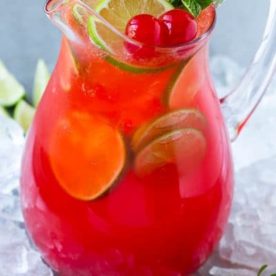 A glass pitcher of cherry limeade garnished with slices of fresh lime and maraschino cherries.