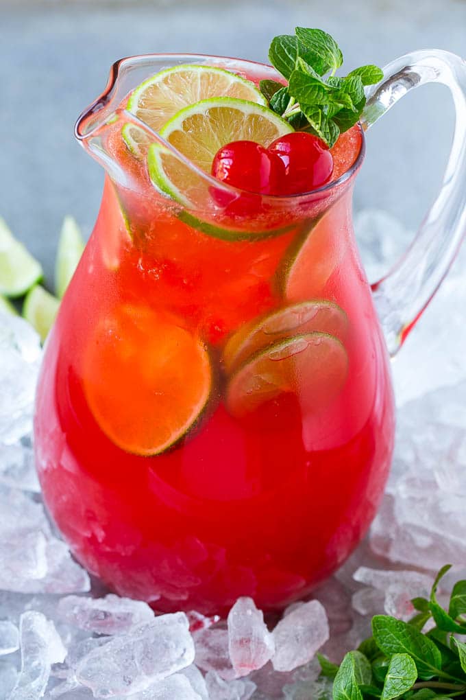 A glass pitcher of cherry limeade garnished with slices of fresh lime and maraschino cherries.