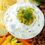 A bowl of dill pickle dip topped with chopped pickles and surrounded by vegetables, pretzels and potato chips.