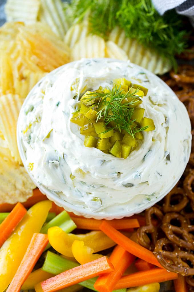 A bowl of dill pickle dip topped with chopped pickles and surrounded by vegetables, pretzels and potato chips.