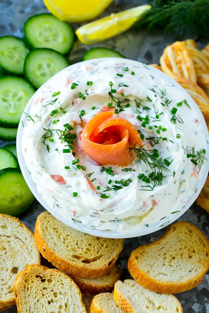 A bowl of smoked salmon dip surrounded by crostini, cucumbers and breadsticks.