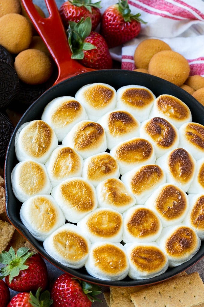 A skillet of s'mores dip with melted chocolate chips and toasted marshmallows, surrounded by graham crackers, cookies and strawberries.