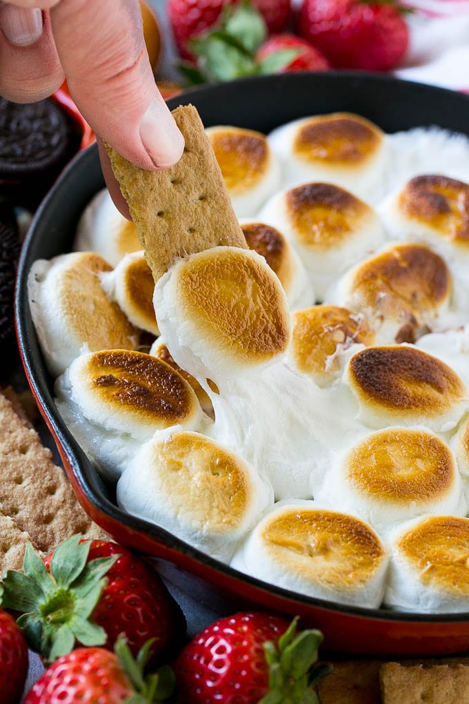 A graham cracker topped with melted marshmallow dipping into a skillet of s'mores dip.