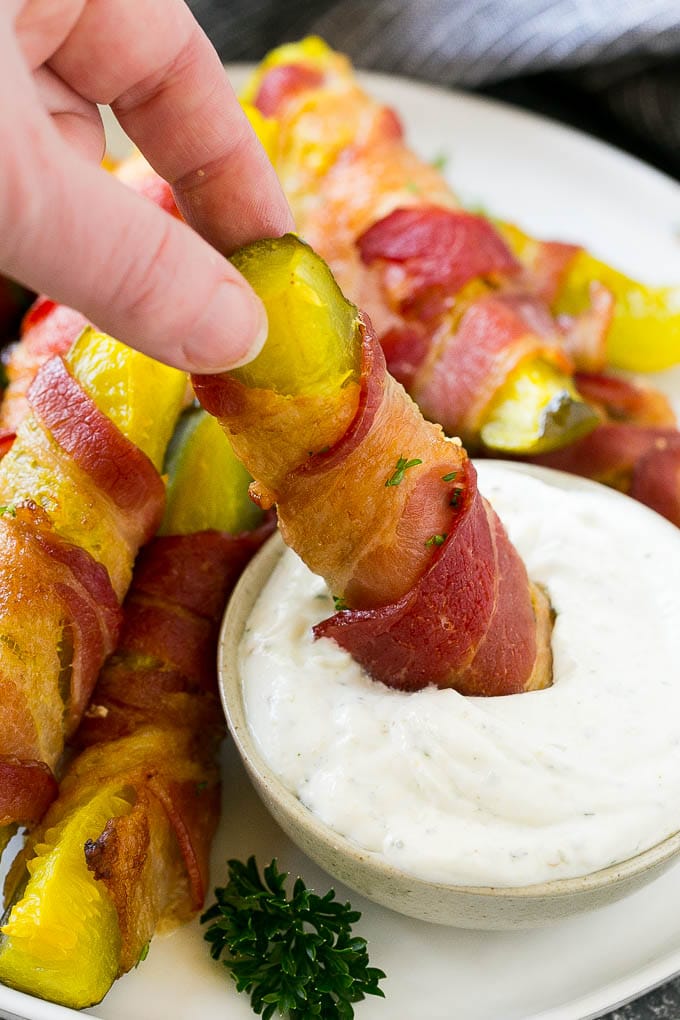 A bacon wrapped pickle being dipped into a bowl of ranch.