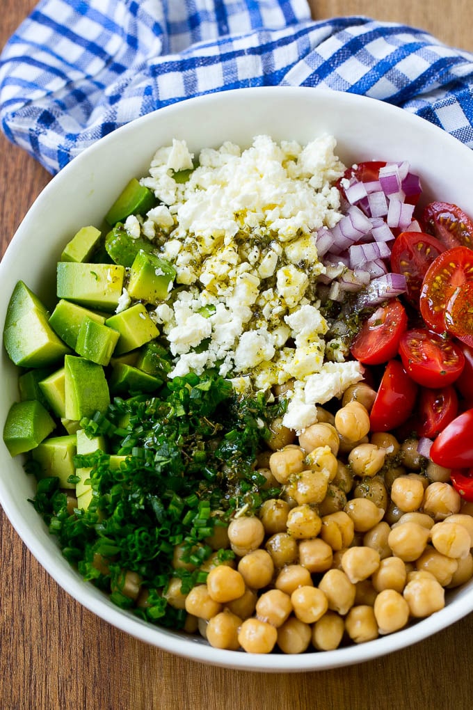 A bowl with chickpeas, chives, avocado, feta, red onion and tomatoes with a Greek dressing drizzled over the vegetables.