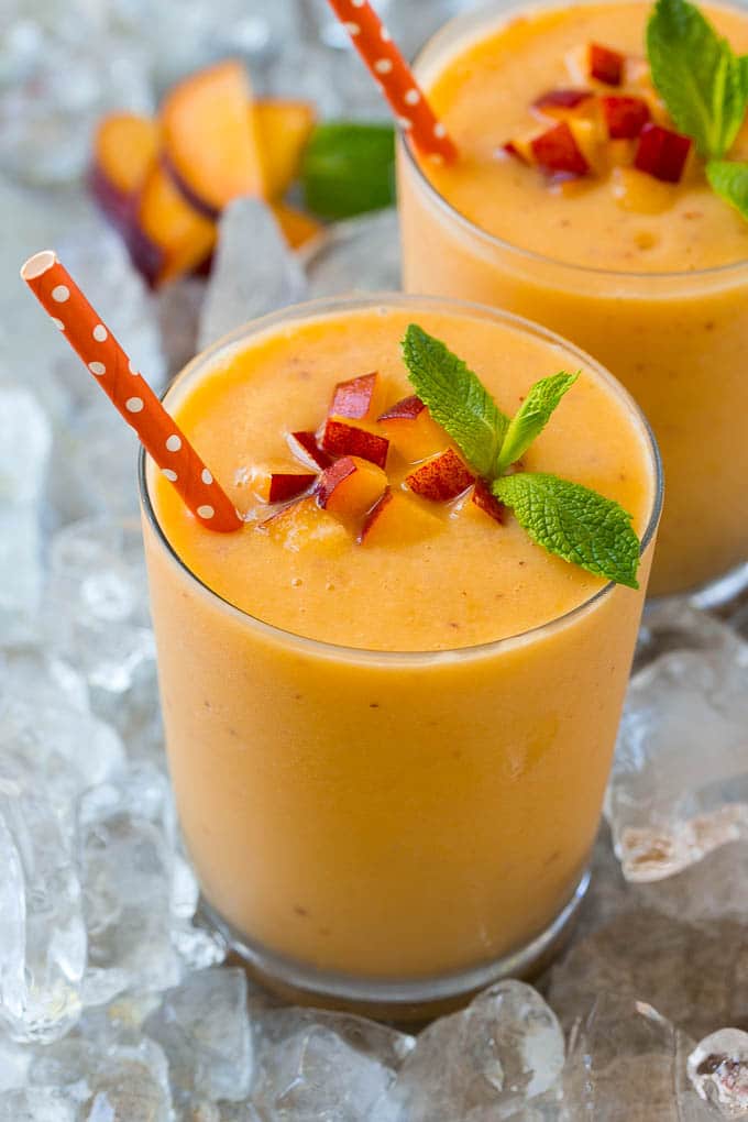 Peach smoothie in a clear glass in a bed of crushed ice, garnished with diced peaches and mint.