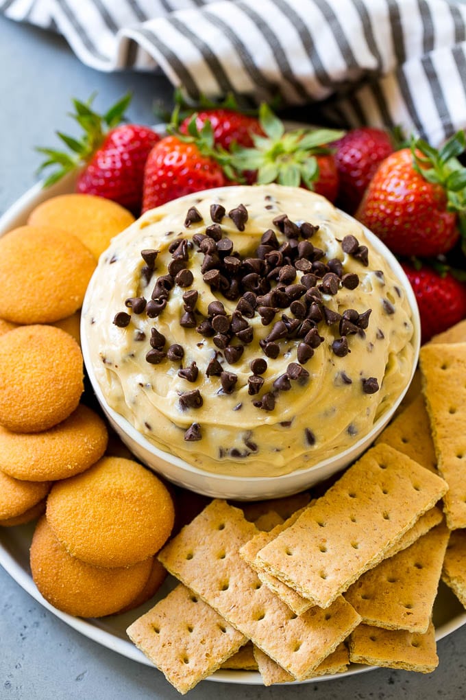 A bowl of cookie dough dip topped with chocolate chips and served with cookies and fruit for dipping.