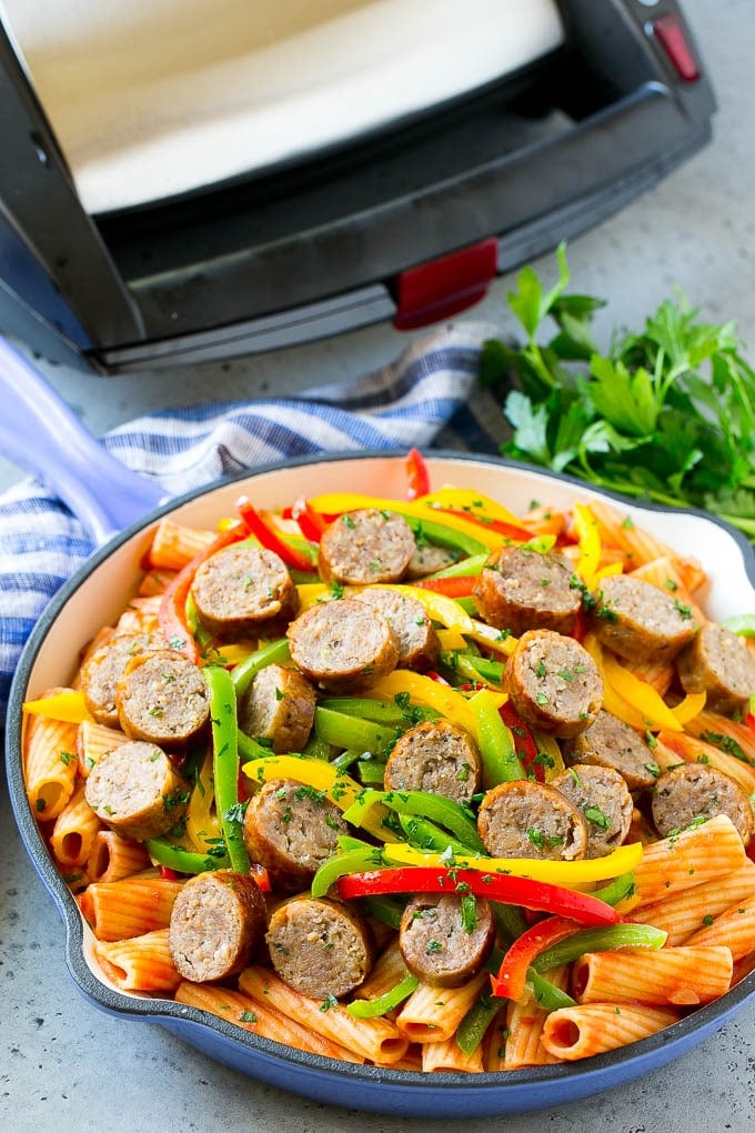 Sausage and pepper pasta in a skillet with a sausage grill in the background.