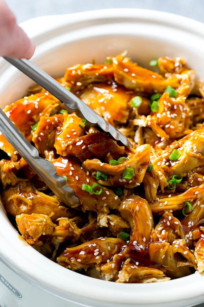 A slow cooker full of teriyaki chicken with tongs in it.