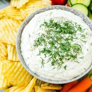 A bowl of creamy dill dip surrounded by potato chips and vegetables.