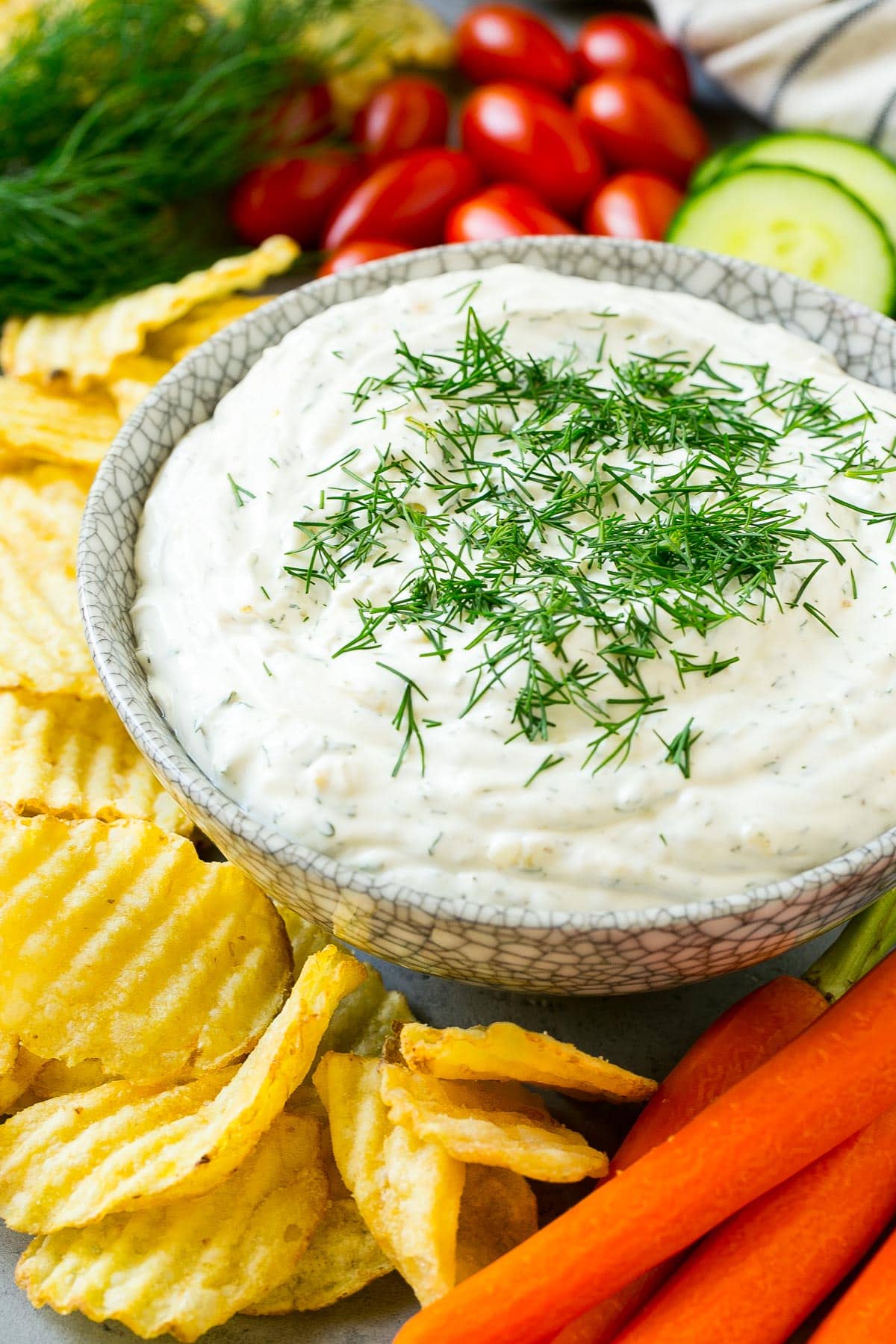 A side view of a bowl of dill dip served with potato chips, carrots and cucumbers.