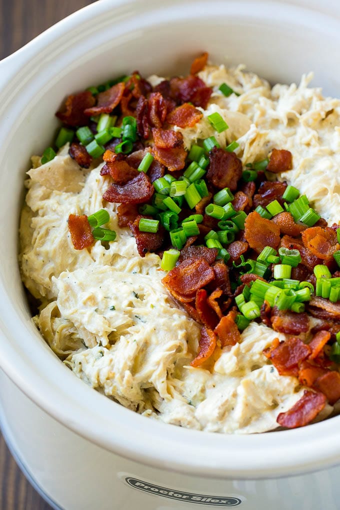 A slow cooker full of shredded crack chicken topped with bacon and sliced green onions.