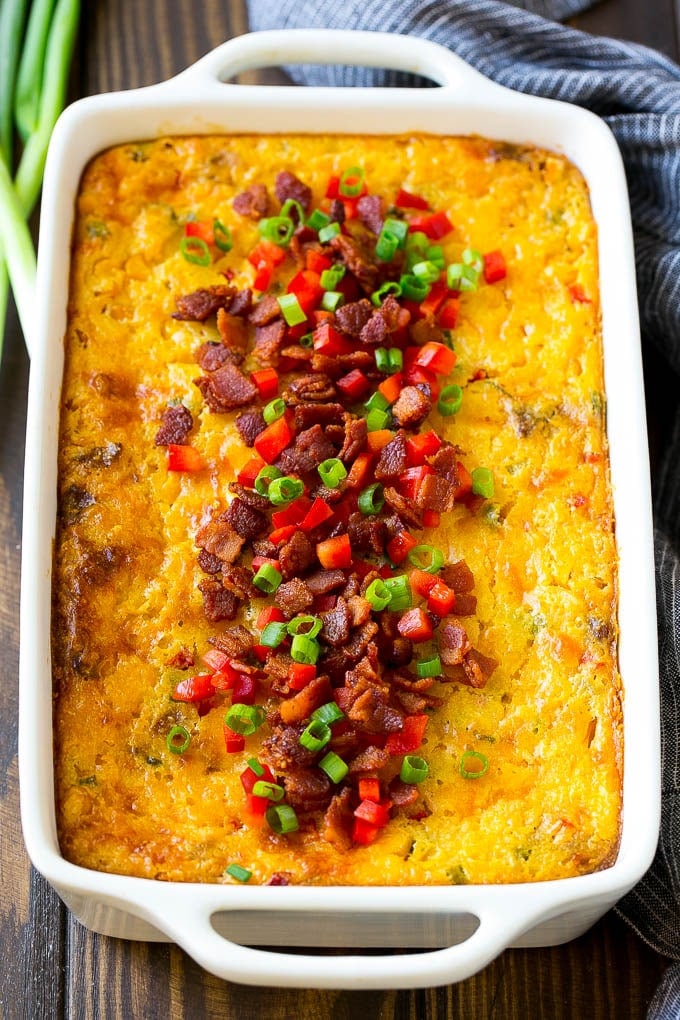 A casserole of loaded corn casserole topped with bacon, red peppers and green onions.