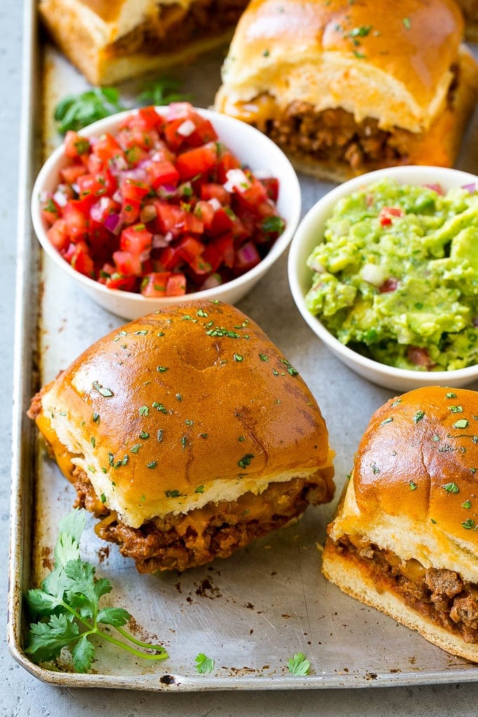 A tray of taco sliders filled with meat and cheese, then served with guacamole and salsa.