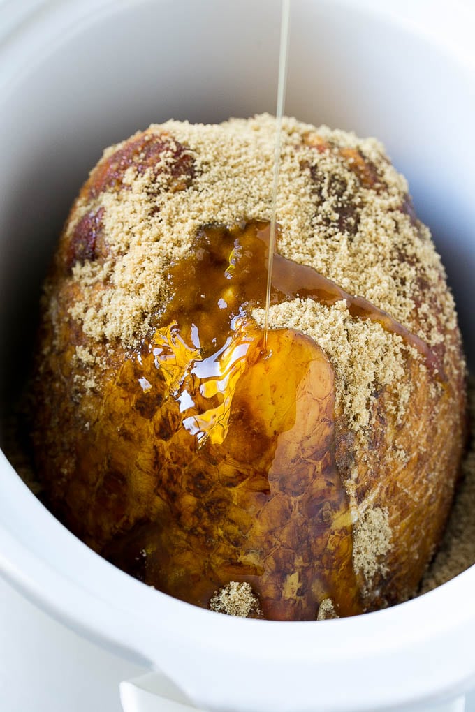 A crock pot ham with maple syrup being poured over the top.