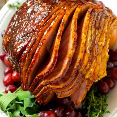 A brown sugar glazed smoked ham on a serving platter surrounded by grapes and fresh herbs.