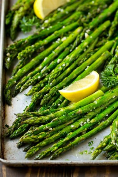 Roasted asparagus on a sheet pan, topped with chopped parsley and garnished with lemon wedges.