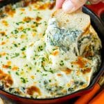 A piece of bread scooping into a pan of spinach dip.