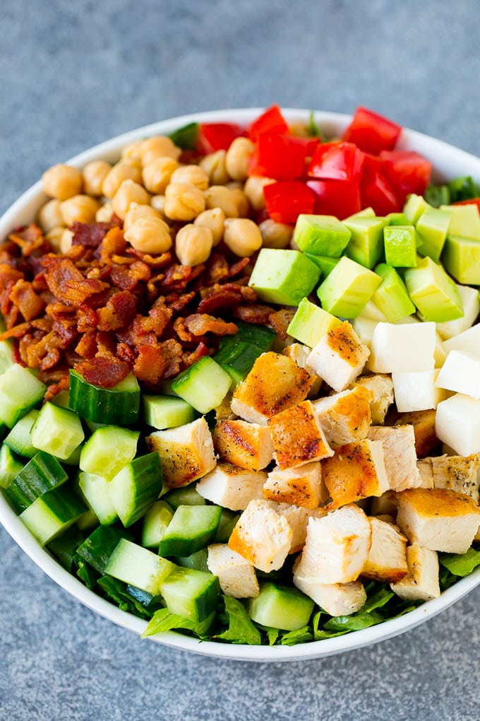 A bowl of lettuce topped with chopped chicken, cucumber, avocado, bacon, chickpeas and tomatoes.