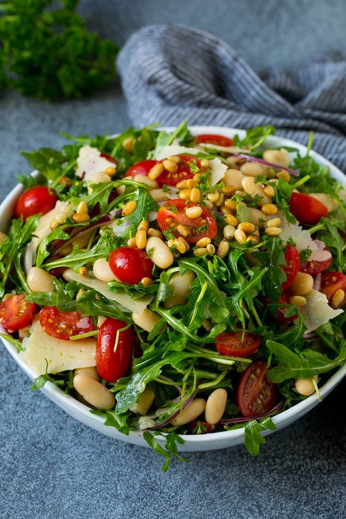 A bowl of arugula salad with cherry tomatoes, pine nuts, red onion and shaved cheese.