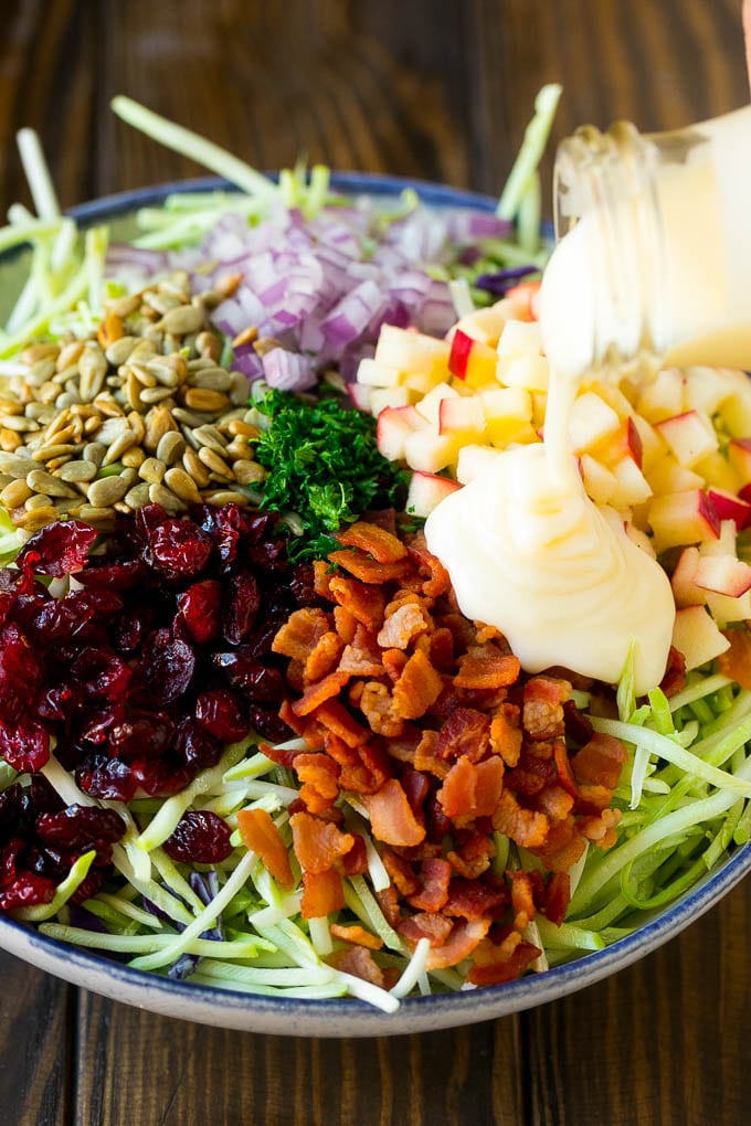 Creamy dressing being poured over broccoli slaw, bacon and apples.