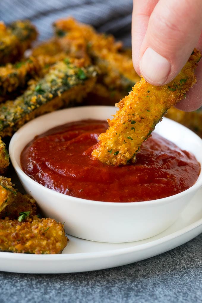 A hand dipping a piece of fried zucchini into marinara sauce.